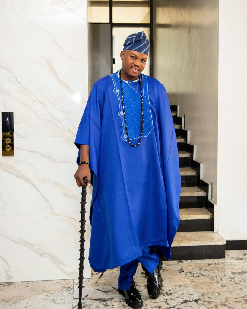 Portrait of a man in vibrant blue traditional attire, standing gracefully indoors with a walking cane.